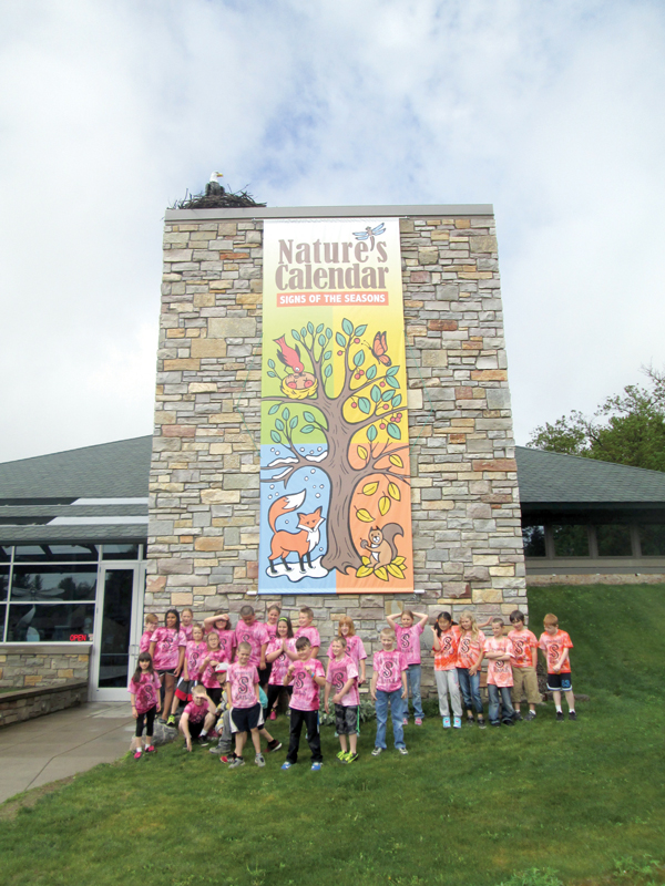 Third graders from Spooner, WI, pose for a class photo under our exhibit banner after learning all about the phenology of hummingbirds, frogs, and weasels. Photo by Emily Stone.